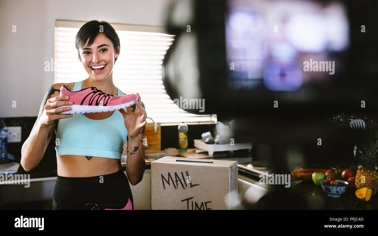 Social media influencer reviewing sports shoe. Smiling young woman vlogging about women's sports shoe and filming herself at home on a video camera. Stock Photo