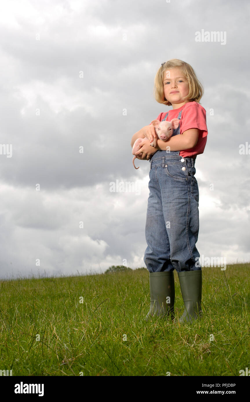 Girl in wellington boots standing in field and holding piglet Stock