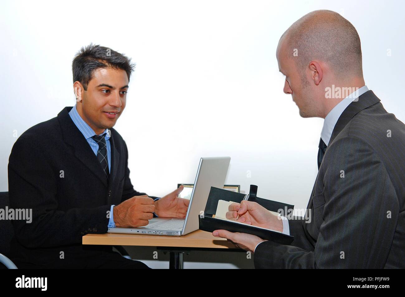 Man sitting with a laptop on desk in front of him, another man sitting opposite taking notes Stock Photo