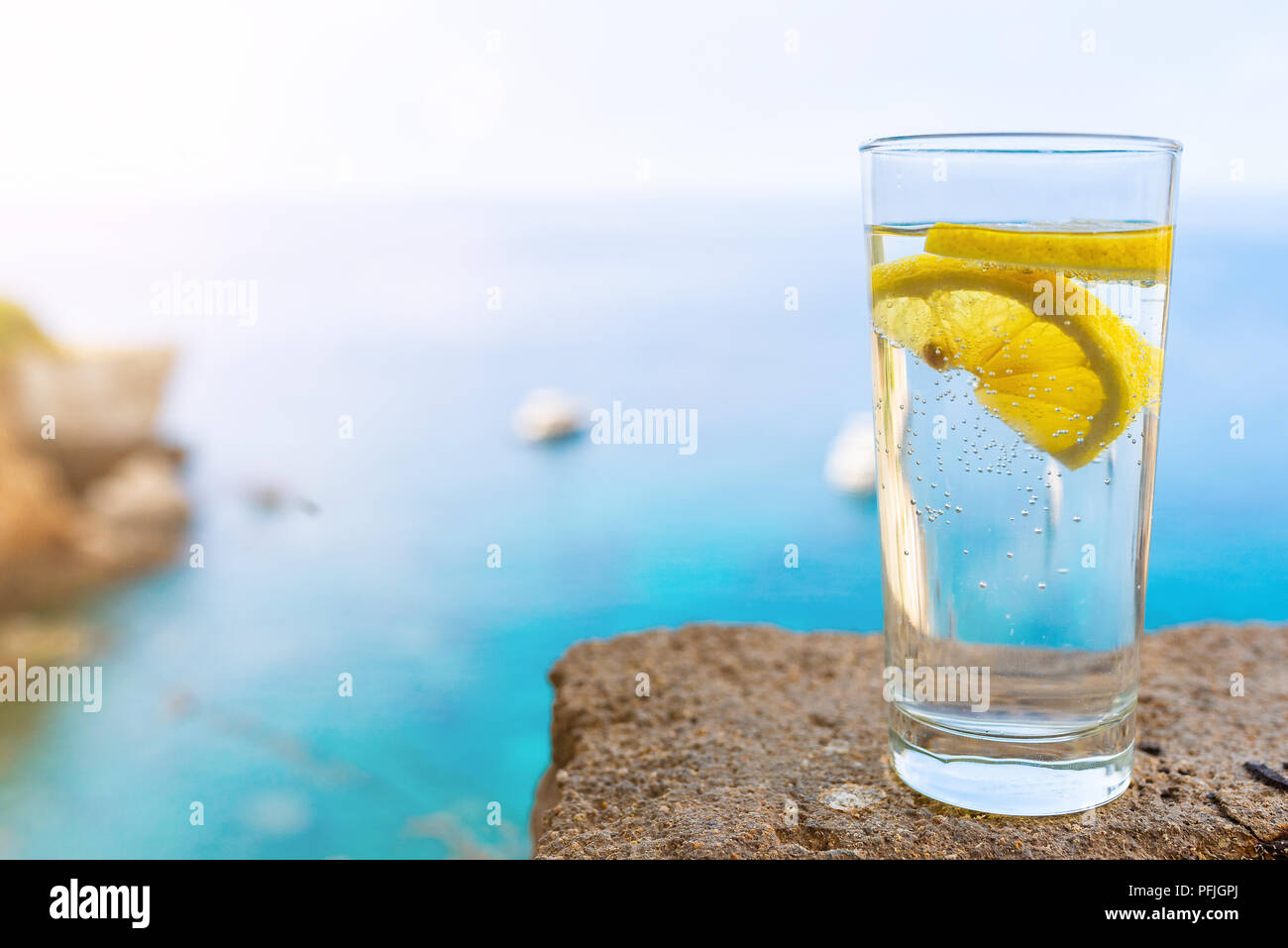 drinking glass with cold carbonated water or soft drink and lemon slice against blue sea and sky Stock Photo