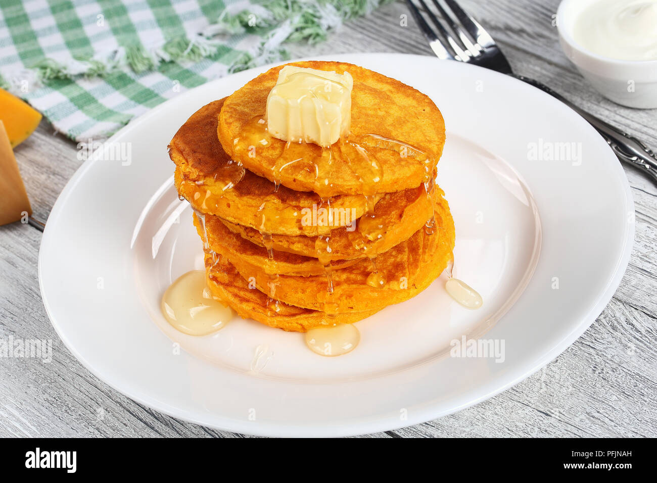 stack of pumpkin pancakes topped with piece of butter and pour over with honey on white plate on kitchen tablecloth, view from above Stock Photo