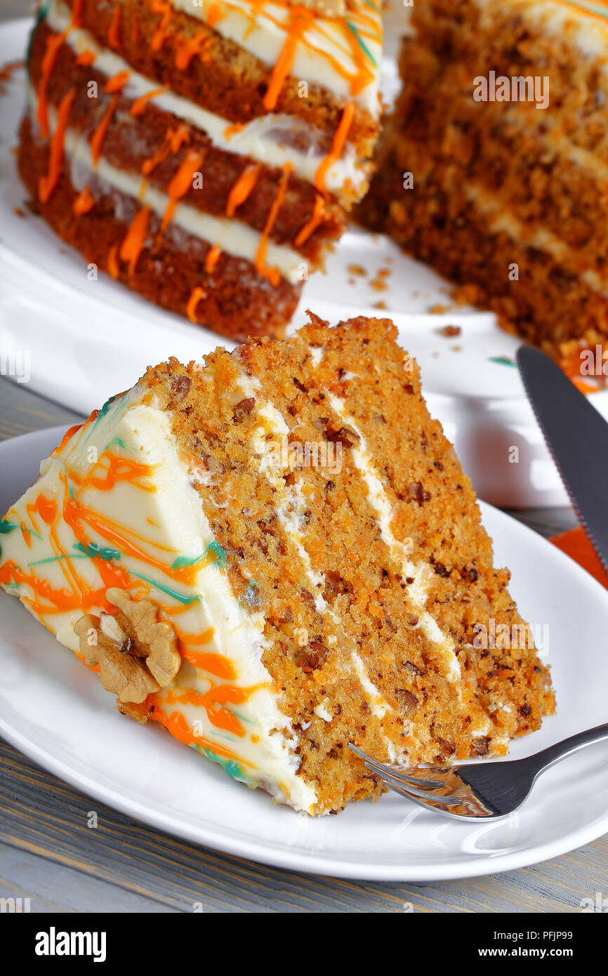 piece of classic carrot cake with cream cheese frosting decorated with walnuts and drizzled with colorful ganache with whole cake on background, verti Stock Photo