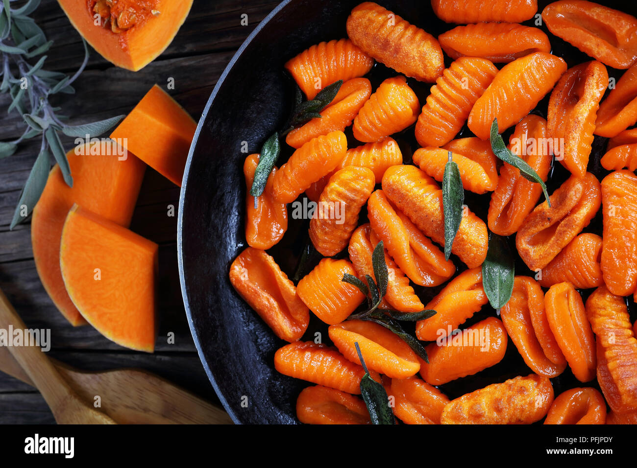 tasty pumpkin gnocchi fried in butter with fresh sage leaves in a skillet. pieces of pumpkin and wooden spatulas on old dark wooden table, horizontal  Stock Photo