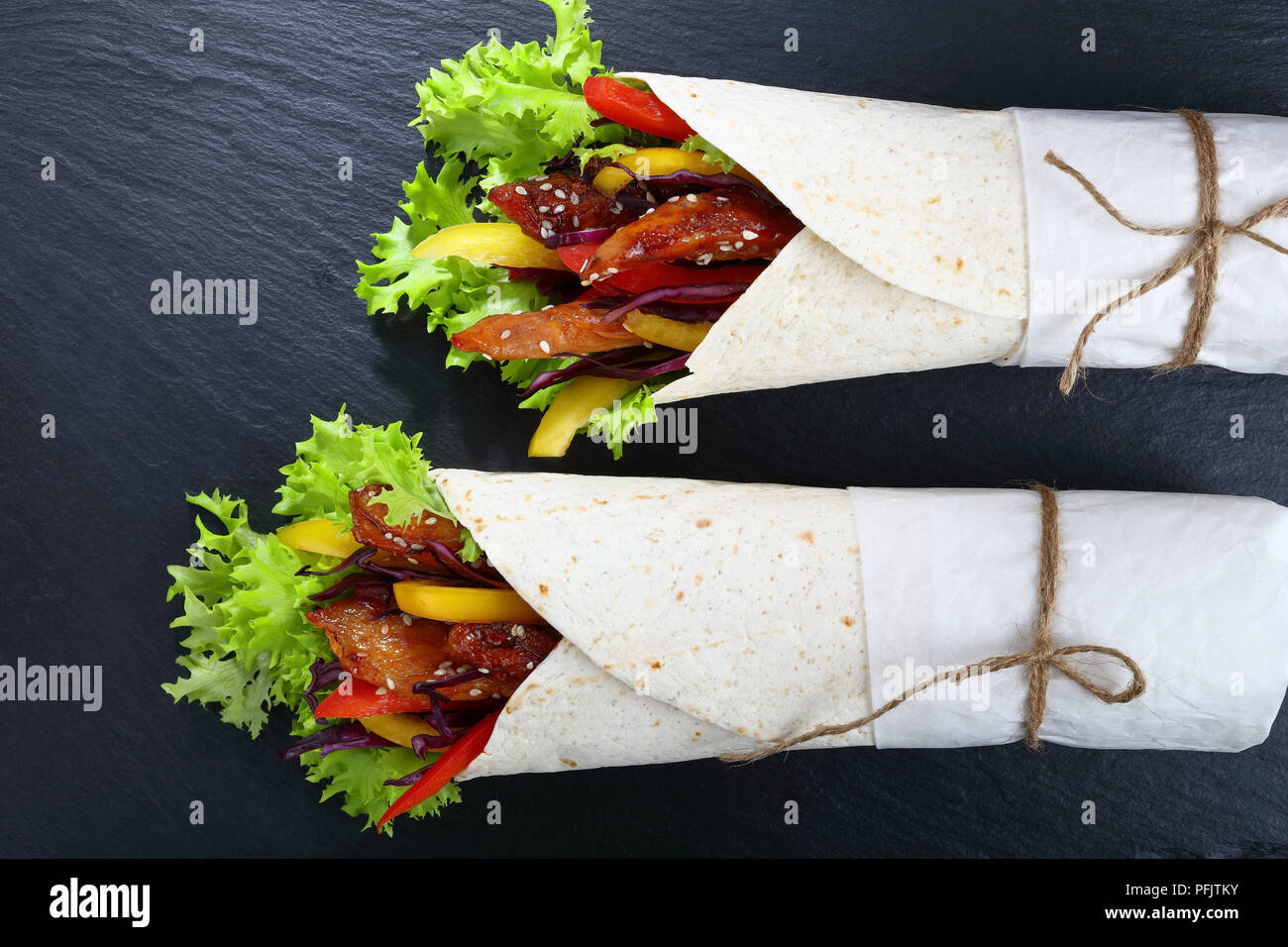 close-up of delicious fresh juicy flatbread sandwich wraps with frisee lettuce, sweet peppers, coleslaw and fried chicken meats on black stone tray, v Stock Photo
