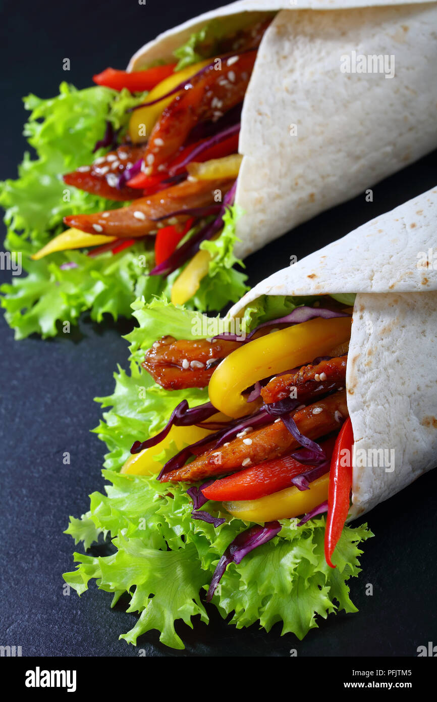 close-up of delicious fresh juicy flatbread sandwich wraps with frisee lettuce vegetables salad and  fried spicy chicken fillet on black stone tray, v Stock Photo