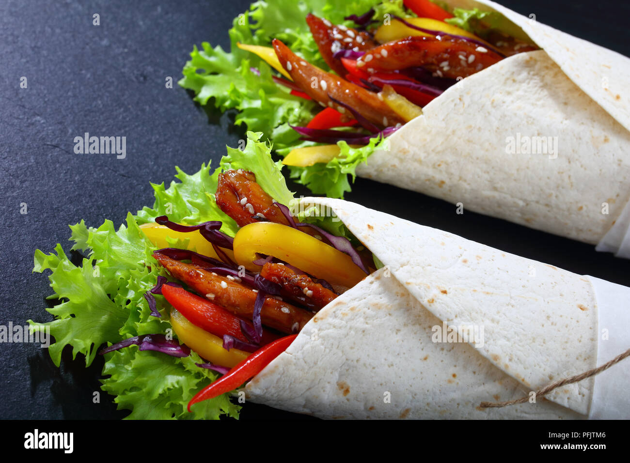 close-up of delicious fresh juicy flatbread sandwich wraps with frisee lettuce vegetables salad and  fried spicy chicken fillet on black stone tray, s Stock Photo