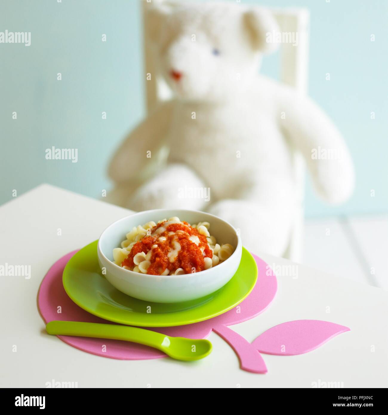 Bowl of pasta with tomato and cheese sauce on table, teddy bear sitting on chair in background Stock Photo