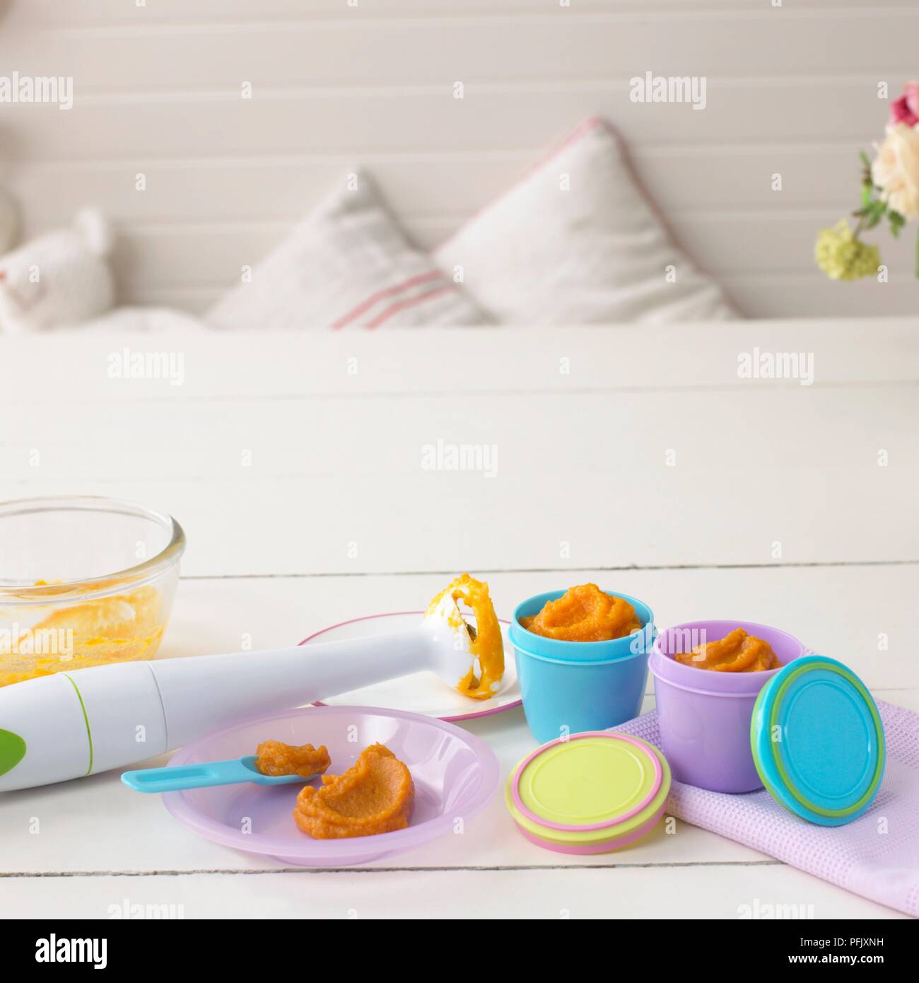 Handheld blender resting on a plate next to plastic pots and a bowl containing carrot puree, on a table Stock Photo
