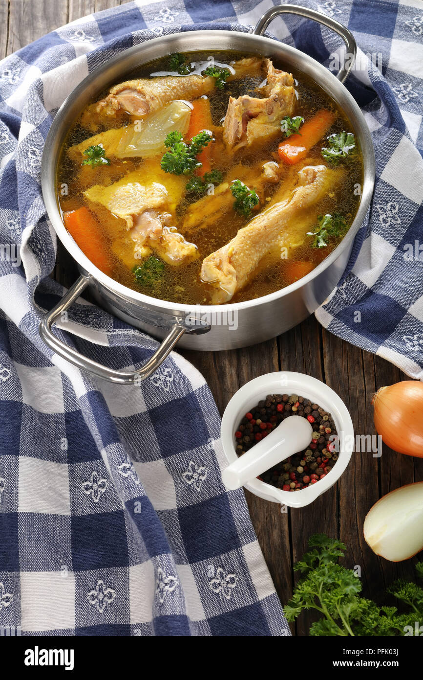 clear Chicken soup with pieces of rooster meat on bone and vegetables in a metal casserole with kitchen towel and ingredients on dark wooden table, ve Stock Photo