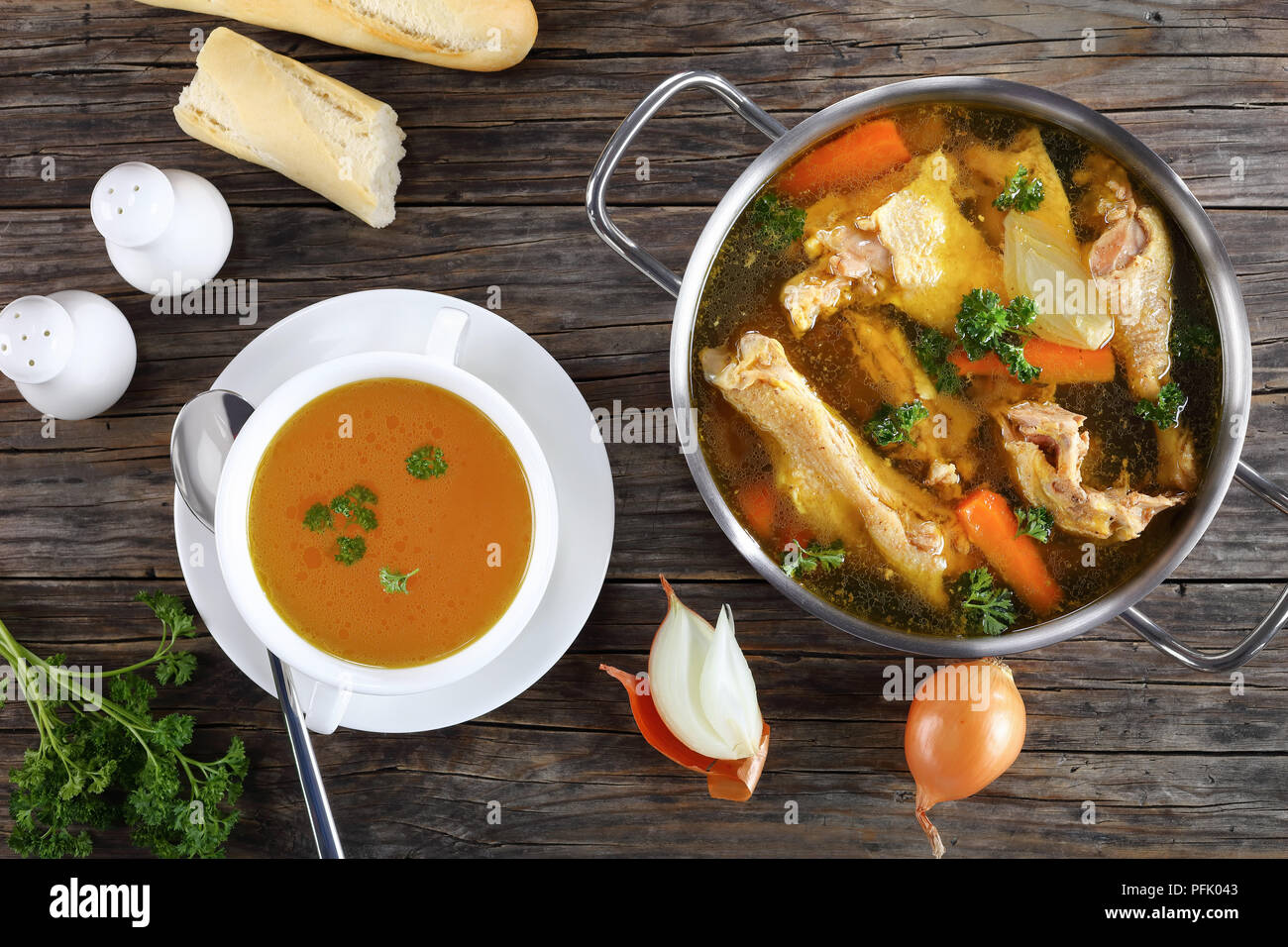 clear Chicken broth with pieces of rooster meat on bone and vegetables in a metal casserole and in a soup cup on dark wooden table, view from above, c Stock Photo