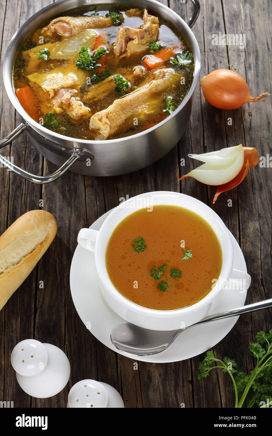 clear Chicken broth with pieces of rooster meat on bone and vegetables in a metal casserole and in a soup cup on dark wooden table, vertical view from Stock Photo