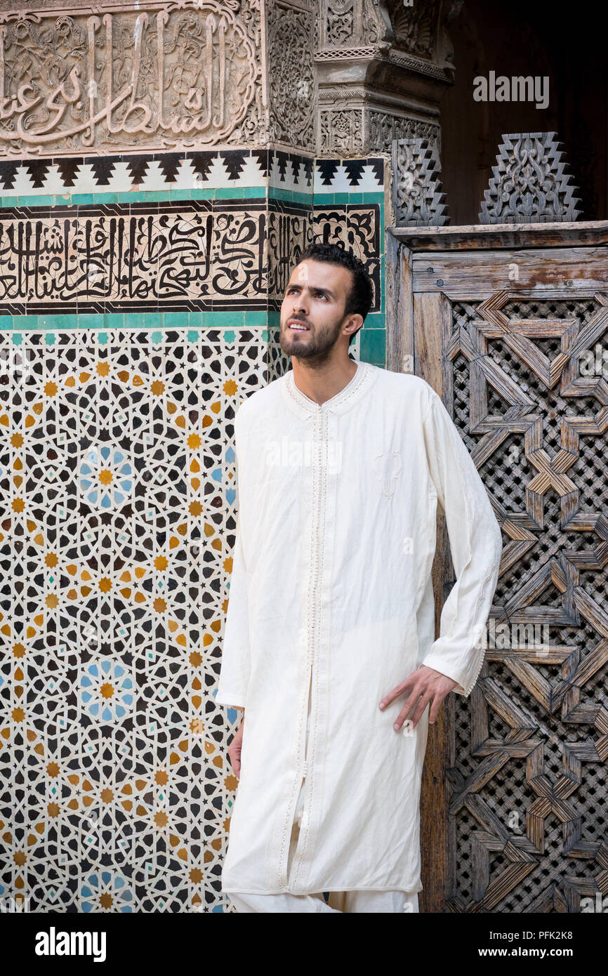 Young muslim man in traditional clothing wearing long white shirt standing in front of the wall with text from Koran and arabesque decoration with flo Stock Photo