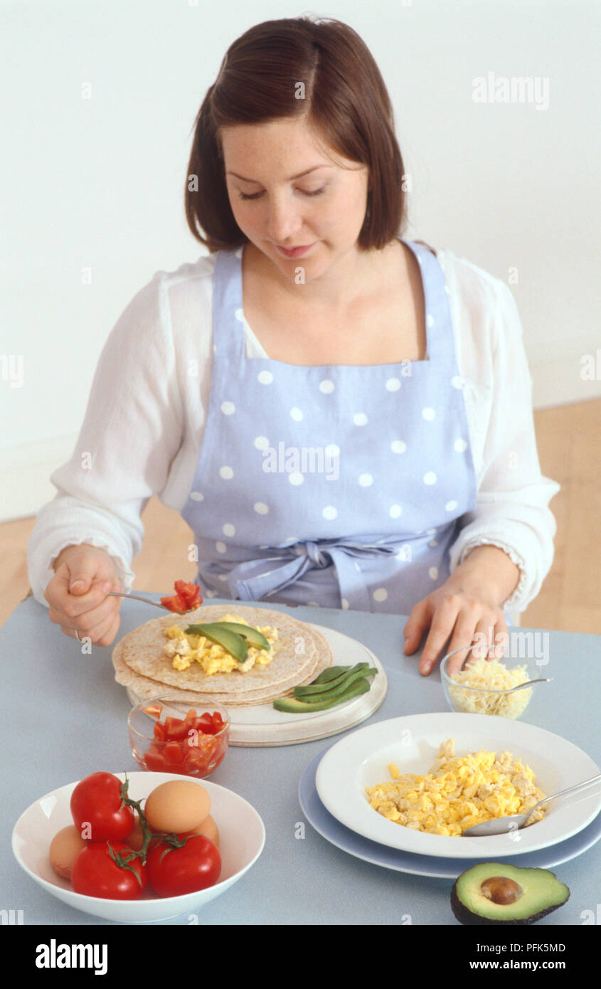 Woman spooning tomatoes on tortilla, on a table with bowls of grated cheese, scrambled eggs, tomatoes, boiled eggs, and a halved avocado Stock Photo