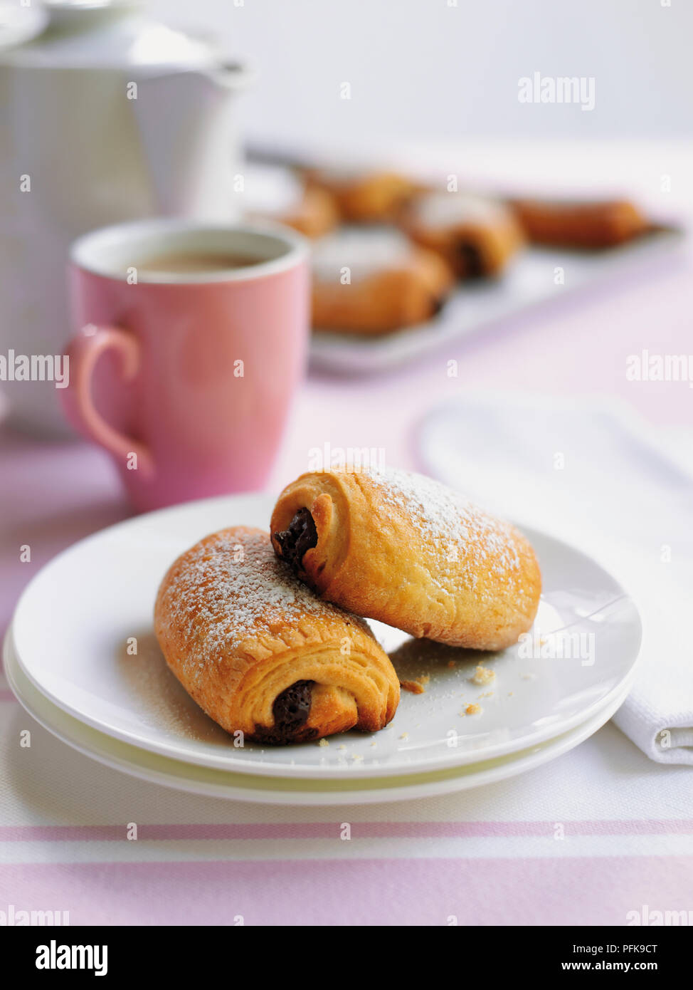 Two pain au chocolat on plates, a mug, coffee pot, and pain au chocolate in background Stock Photo