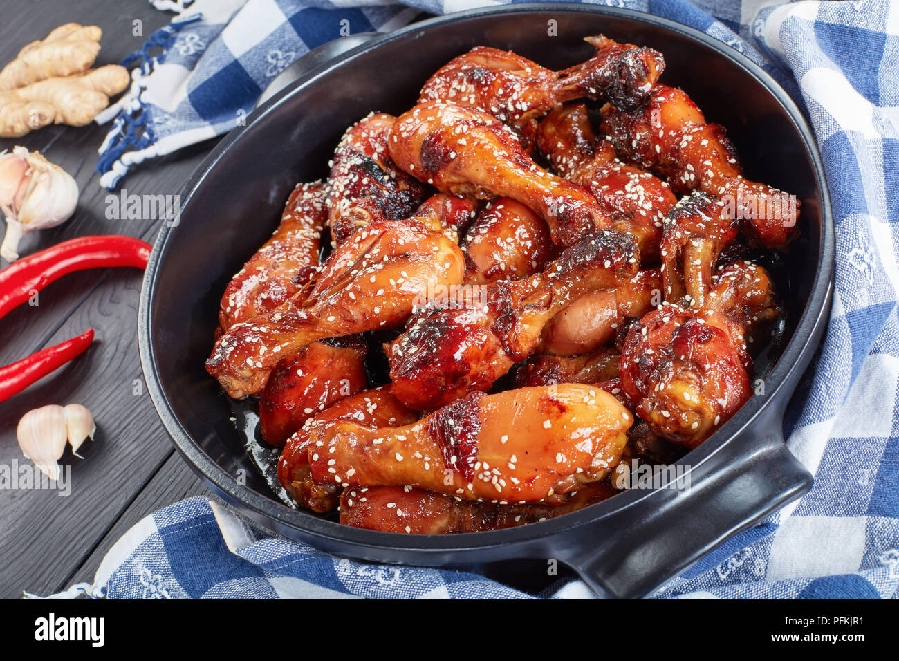close-up of delicious grilled crispy chicken drumsticks sprinkled with sesame seeds in black brazier on wooden table with ingredients for teriyaki sau Stock Photo
