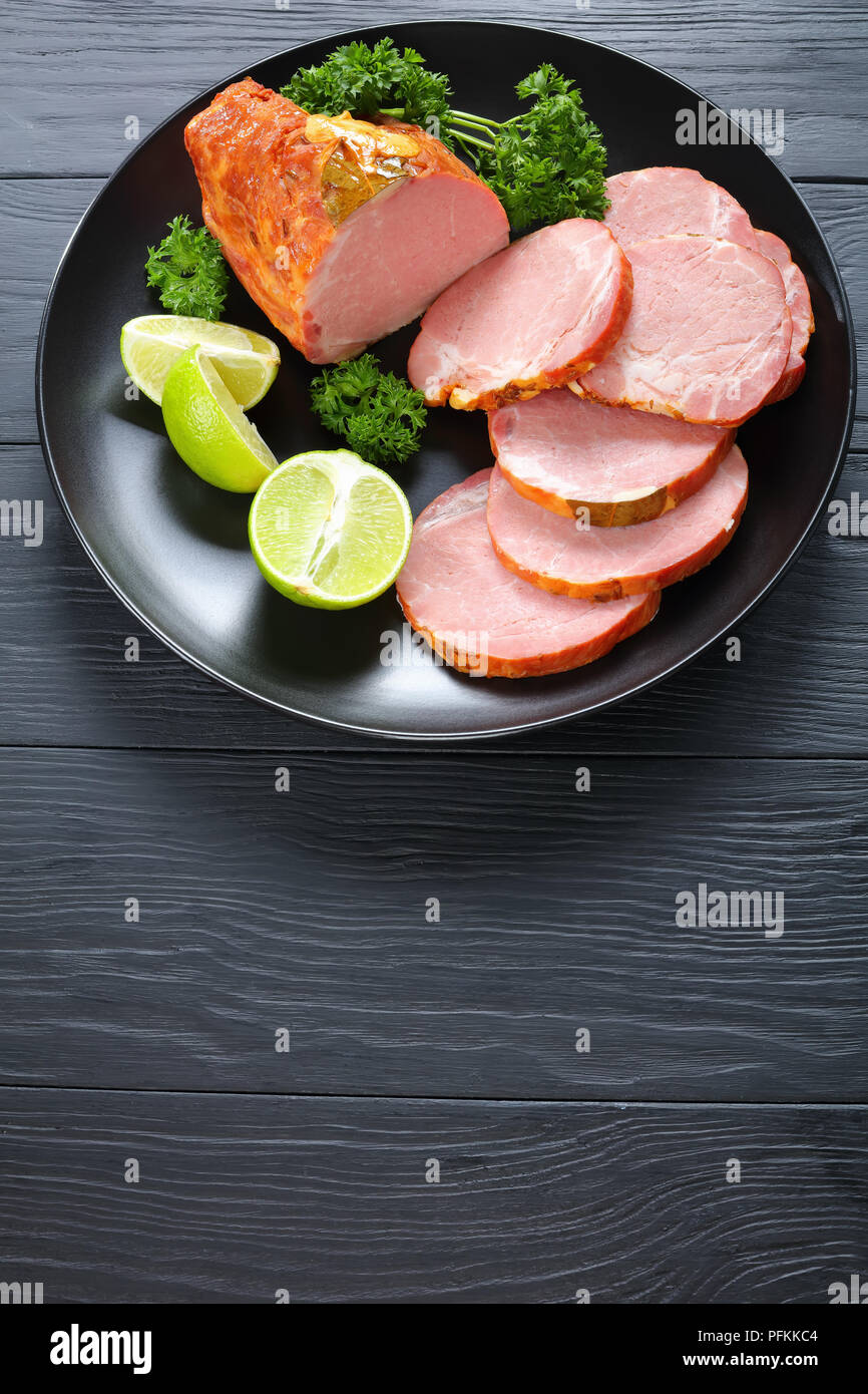 delicious ham cut in slices served with parsley and fresh citrus wedges on black plate on black wooden table, vertical view from above Stock Photo