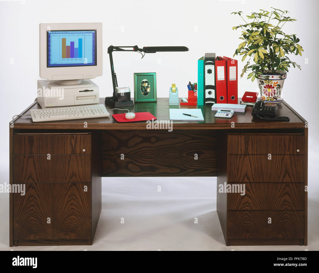 Desk top computer, desk lamp, framed picture, pocket calculator, telephone, various stationery and a houseplant on a wooden desk Stock Photo