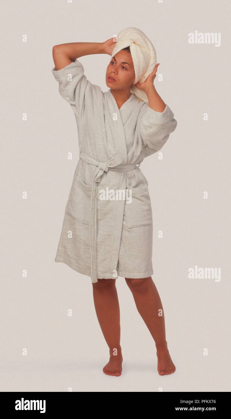 Woman in white bathrobe adjusting towel wrapped around her head, standing, looking away Stock Photo
