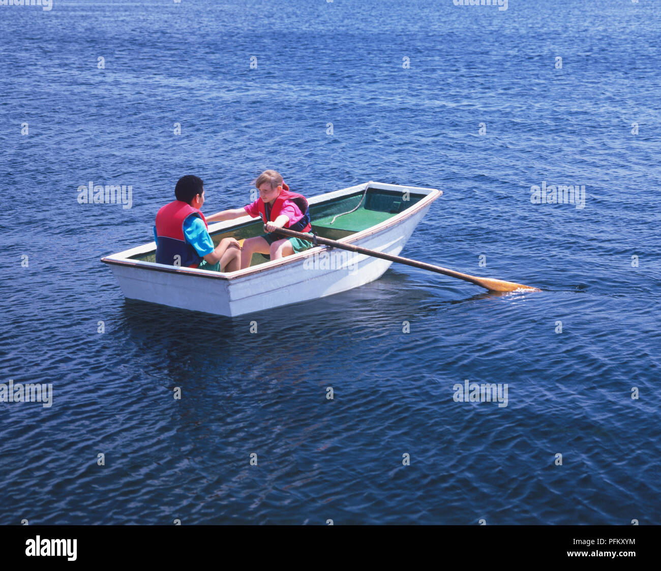 A boy and girl wearing life jackets while rowing a small boat on the open water Stock Photo