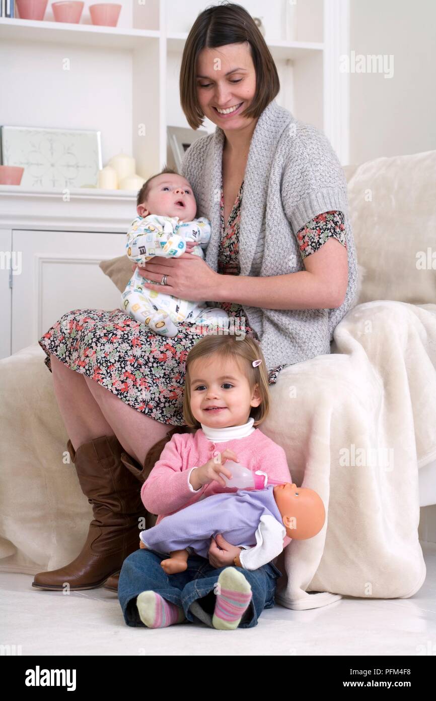 Woman sitting with baby boy on her lap, next to a girl holding plastic  bottle to the mouth of a doll Stock Photo - Alamy