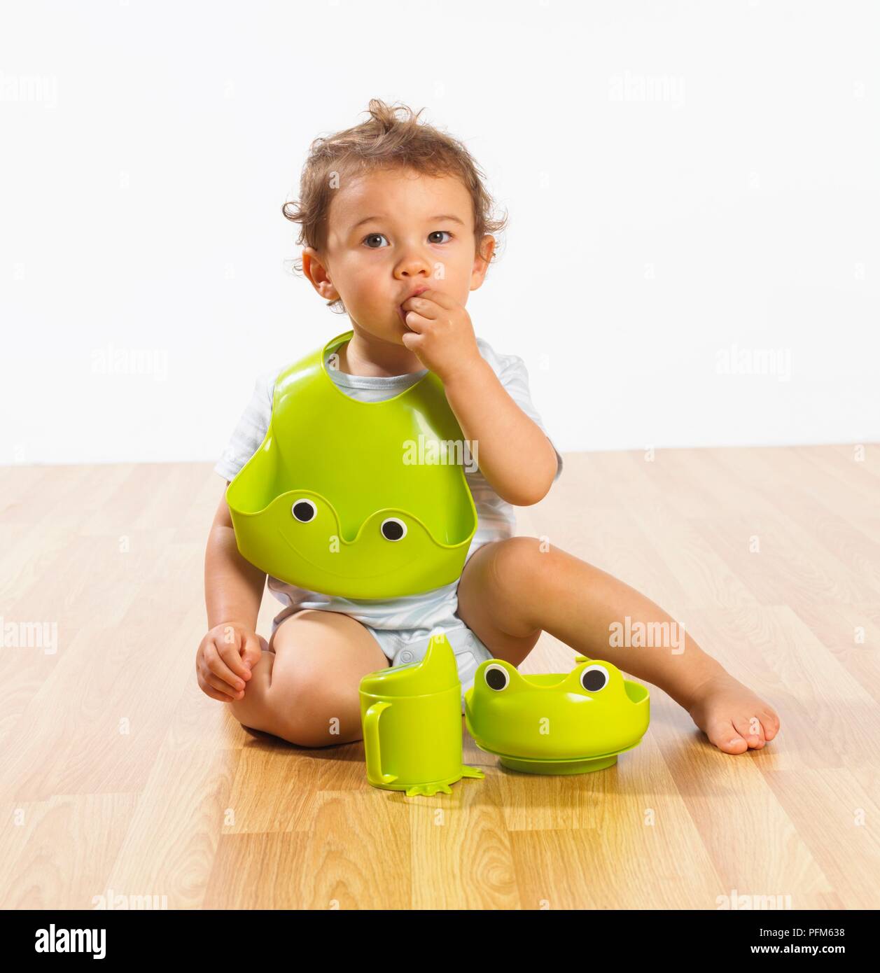 Baby boy with green plastic bib around neck eating with his hand, plastic bowl and mug with spout in front Stock Photo