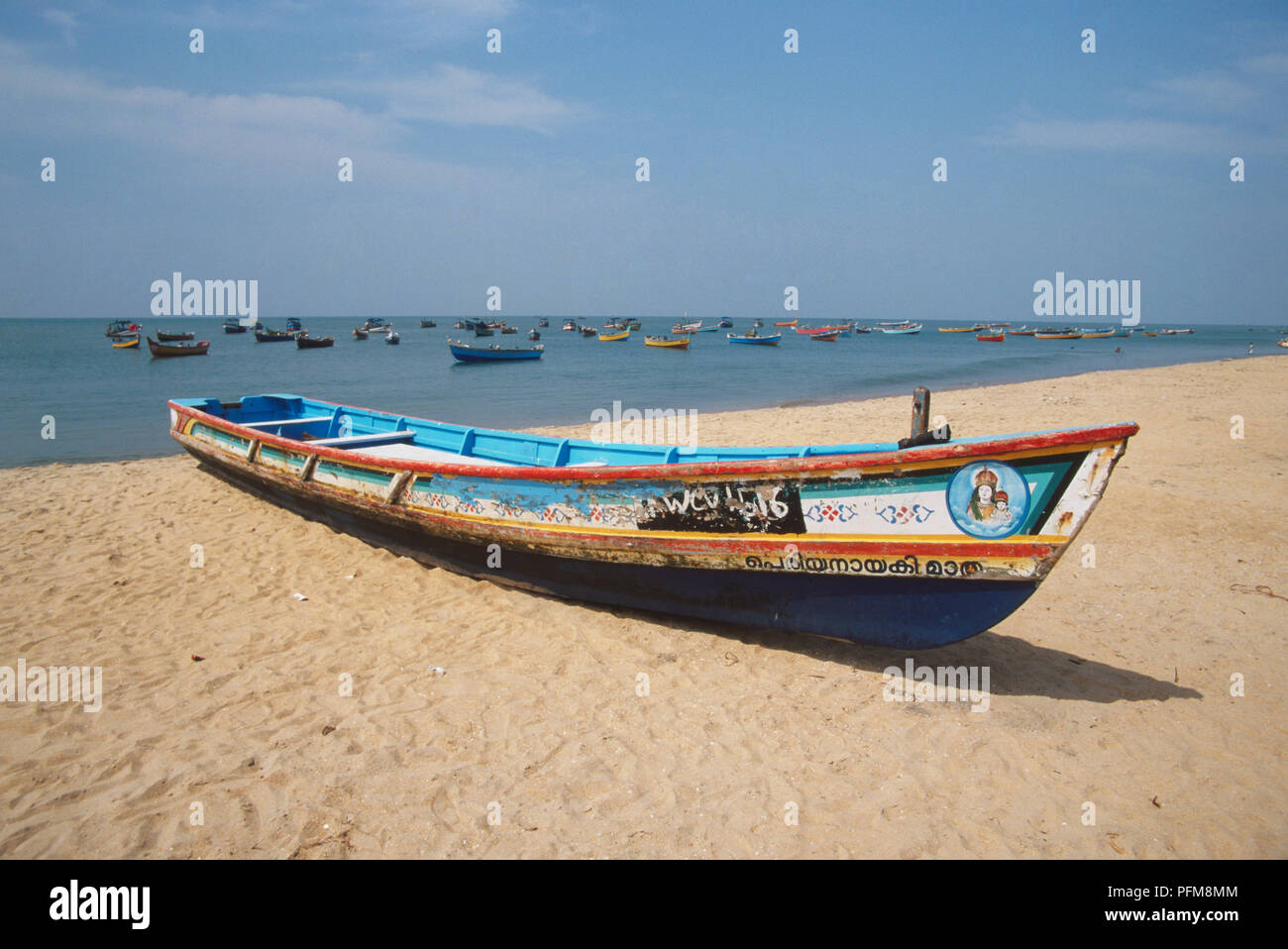 Brightly painted, but weathered, fishing boat on the sands of the Malabar Coast on the Arabian Sea. Many more boats are moored in the sea behind. Stock Photo