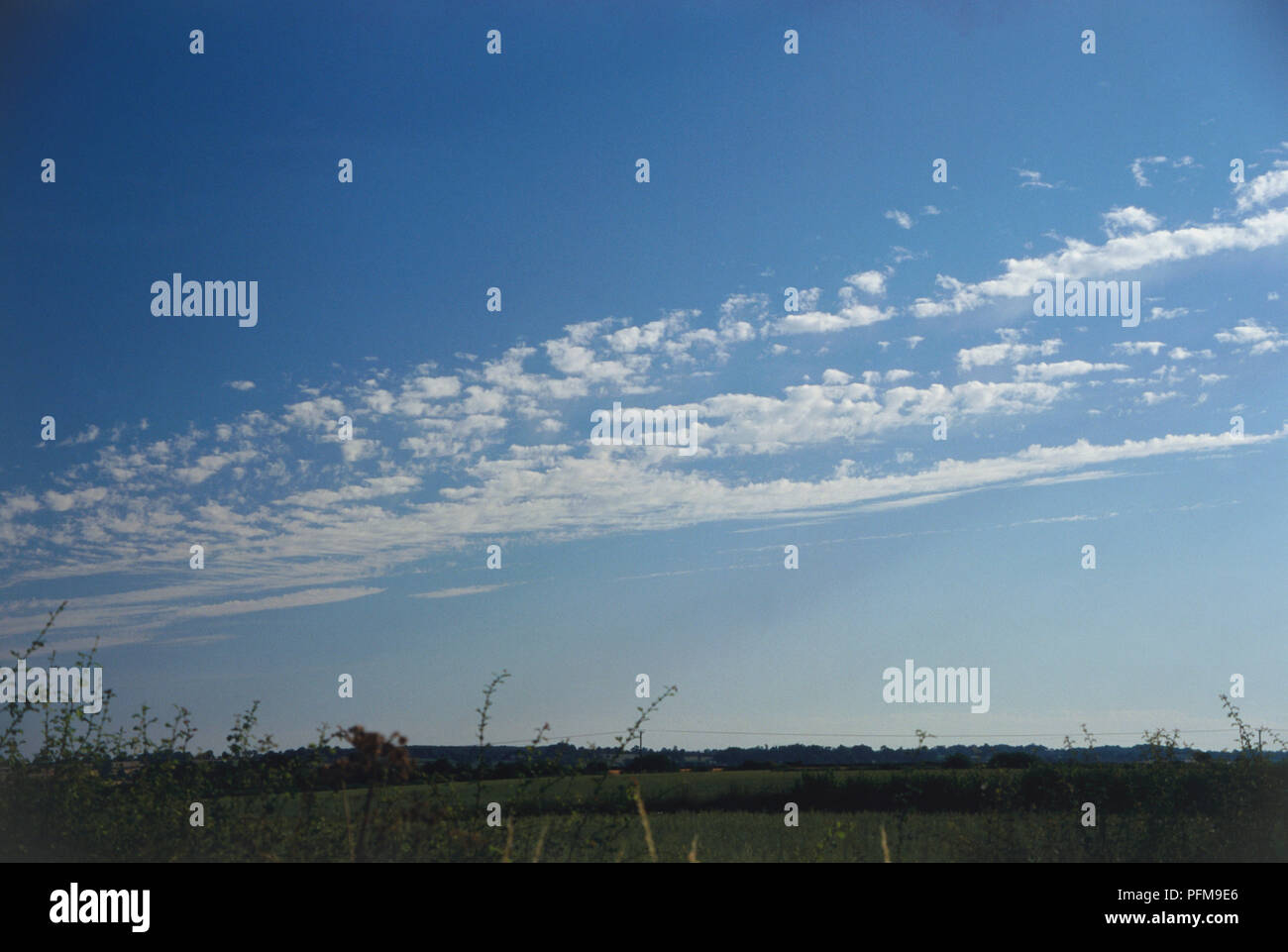 Altocumulus castellanus clouds, thin tufts in parallel lines forming band in blue sky, fields of countryside below. Stock Photo