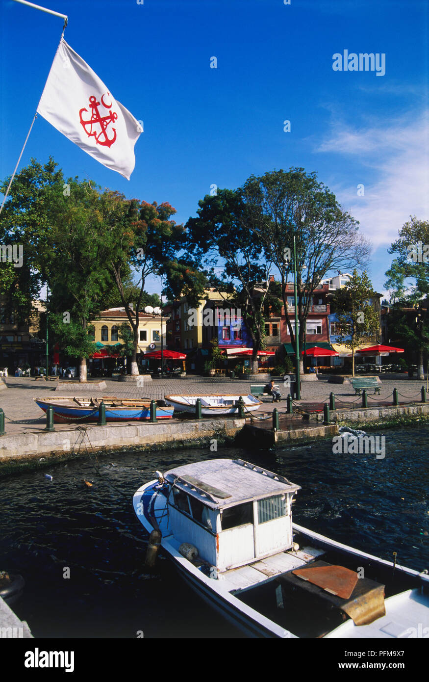 Asia, Turkey, Ortakoy, boat moored to waterfront, square beyond with colourful cafes and tall trees, nautical flag blowing in the breeze. Stock Photo