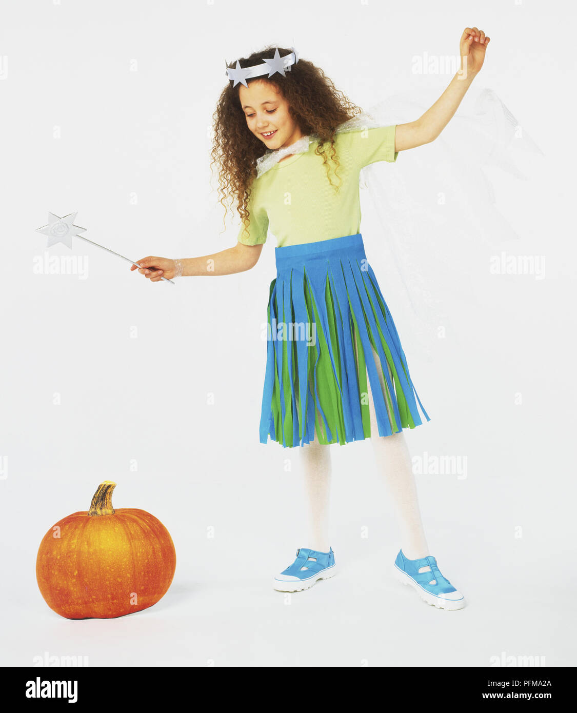 Smiling girl in fairy costume standing and waving wand at pumpkin on the floor, front view Stock Photo