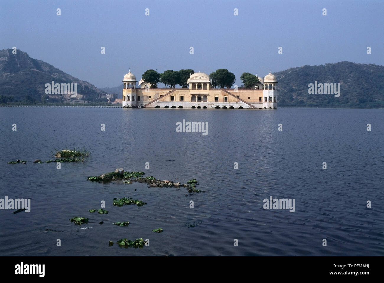 India, Jaipur, picturesque Jal Mahal, Water Palace, seemingly afloat in the monsoon, yellow and white painted palace, semi-octagonal tower topped by elegant cupola at each corner, courtyard treetops visible behind facade, vast lake in foreground. Stock Photo