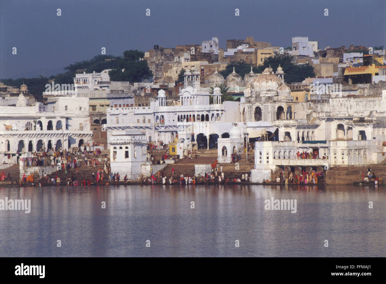 India, Pushkar, ghats beside river, devout Hindu pilgrims bathing in holy ghats to cleanse sins, many people at water's edge reflecting in river, steps leading up to white buildings and mosques on hillside. Stock Photo