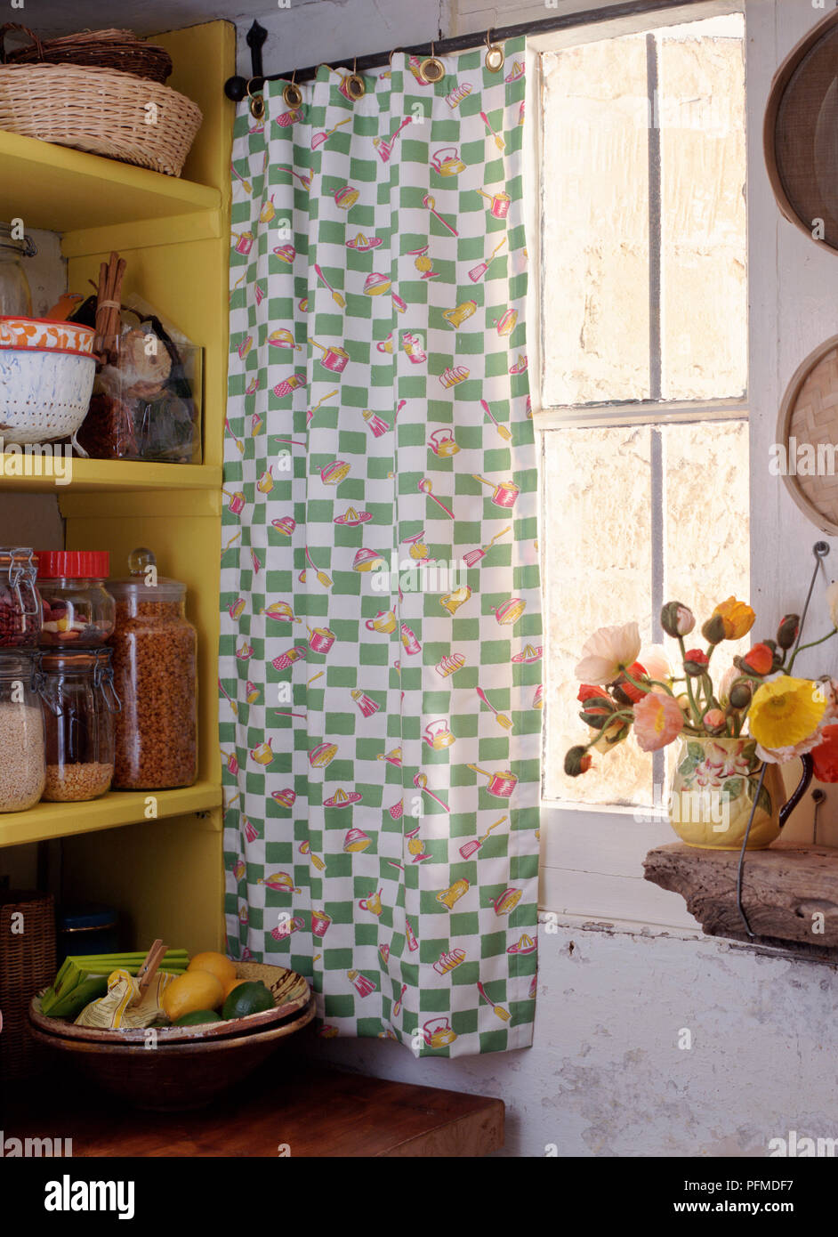Cottage window viewed from inside; eyelet curtain with green check and kitchen utensils on hanging rail over window, vase of flowers standing on rough wooden table, yellow shelves beside. Stock Photo