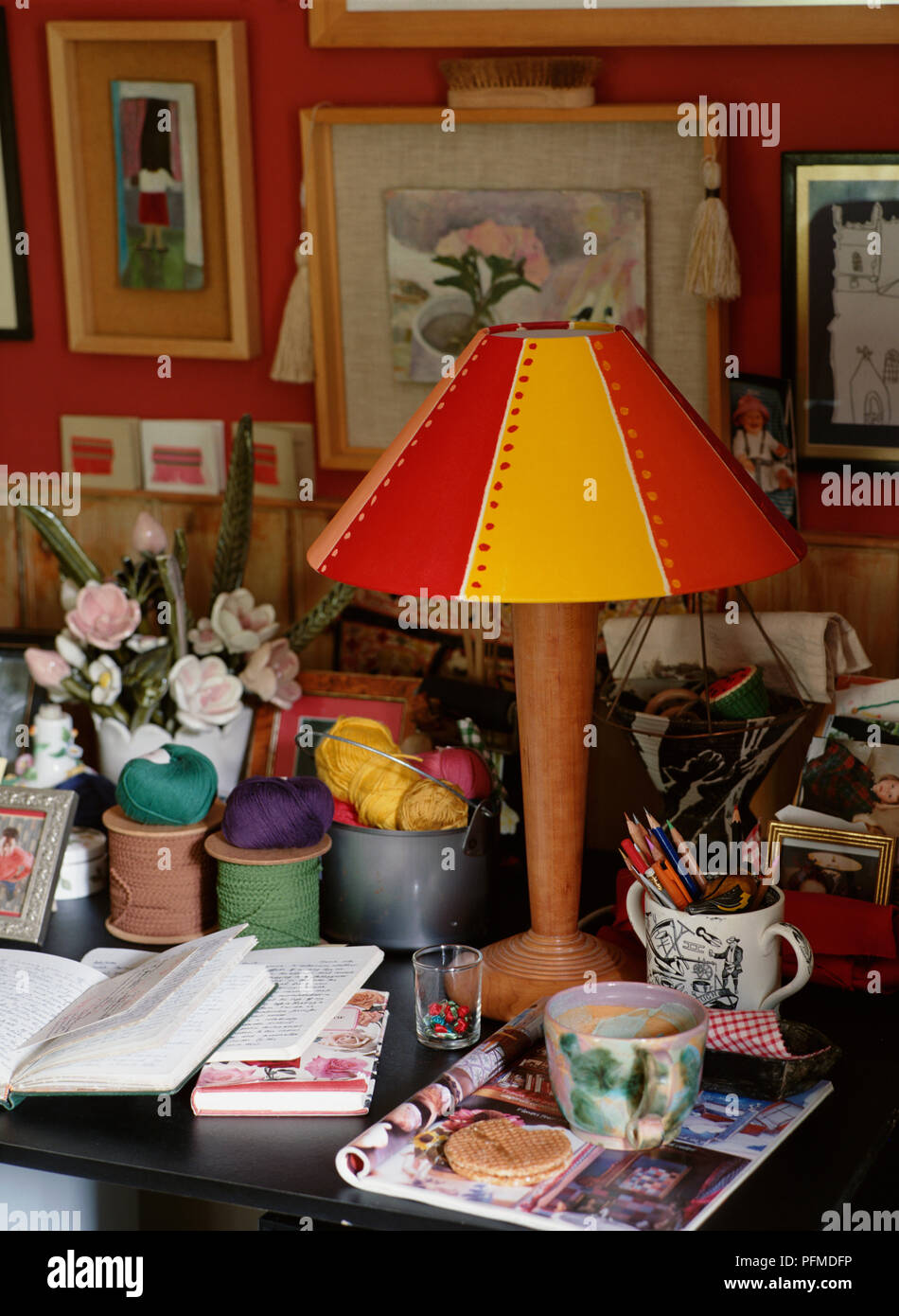 Red and yellow lamp decorated with polka dots, standing on cluttered desk, mugs, books, balls of wool, and vase of flowers surrounding, framed pictures covering red wall in background. Stock Photo