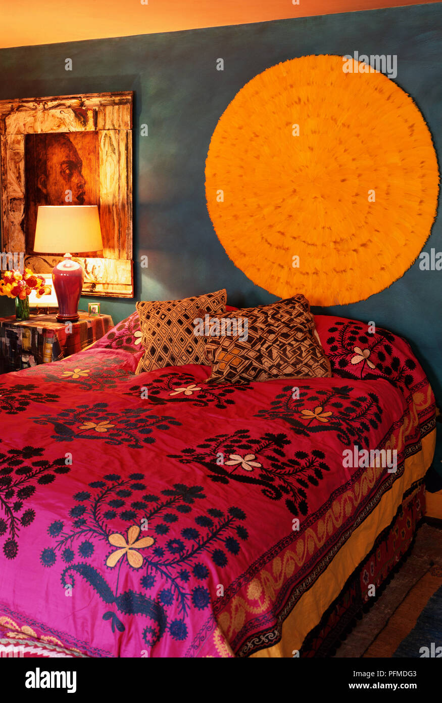 Indian style themed bedroom with pink and red patterned bed cover, gold and brown cushions scattered on bed, dark blue walls, large yellow circular wall decoration, bedside lamp illuminating picture of head in profile. Stock Photo