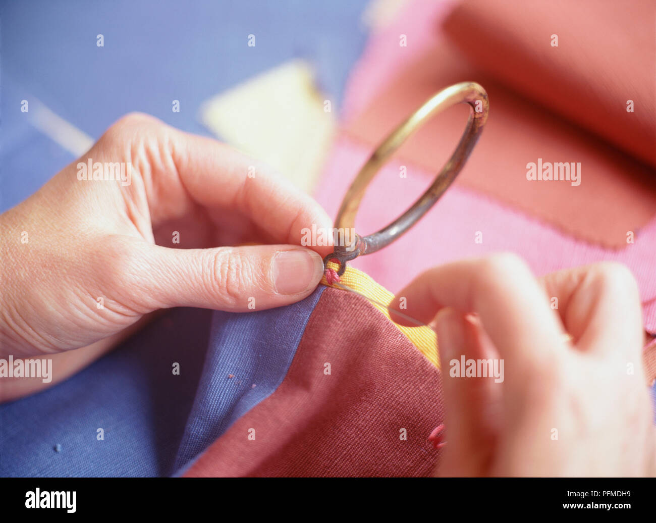 Hands sewing metal curtain rings to red, blue and yellow material, pulling embroidery thread through eyelet rings. Stock Photo