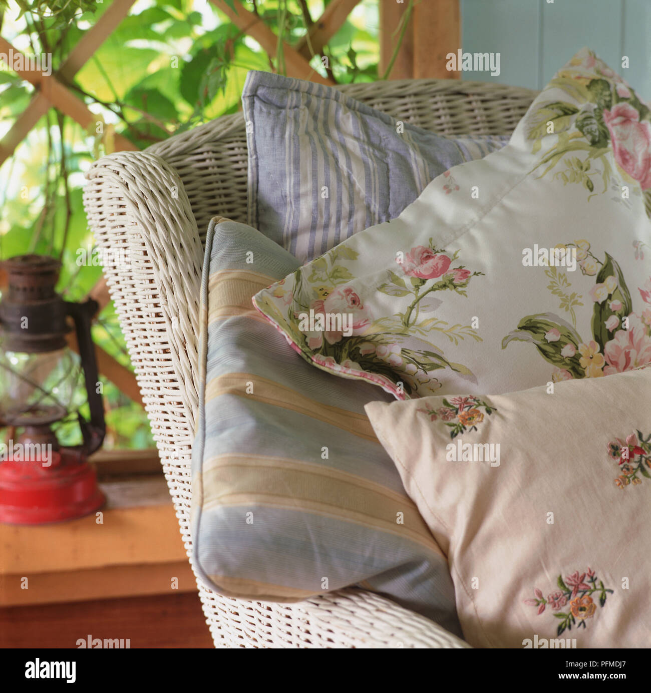 Piles of striped blue and floral white cushions on white wicker chair, wooden trellis and green foliage in background. Stock Photo