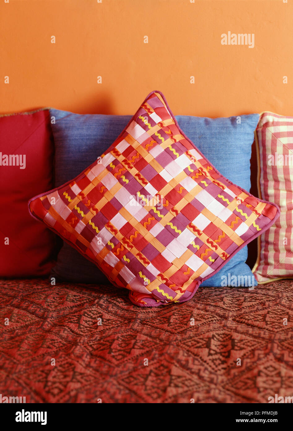 Colourful cushions scattered on red sofa, pink, red and white cushion with woven ribbon cover at front, blue cushion behind, yellow wall in background. Stock Photo