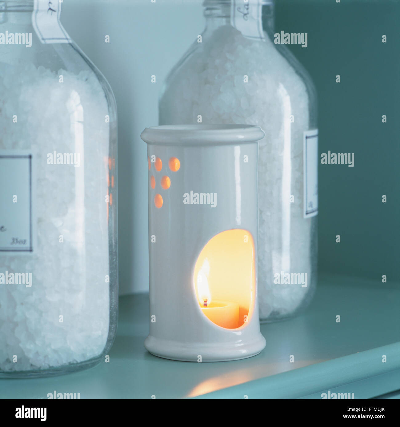Tealight candle with flame burning inside white porcelain oil burner, glass bottles containing salts standing on shelf either side. Stock Photo