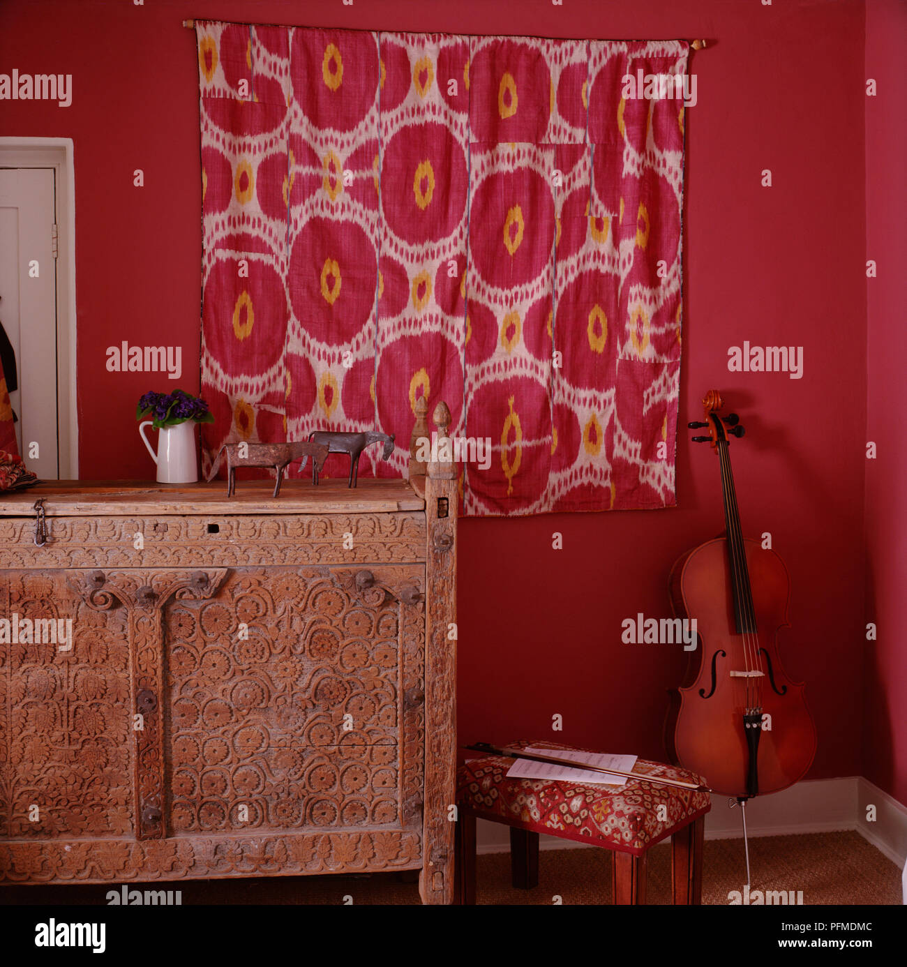 Red room, red and cream fabric hanging over curtain, horse ornaments and jug of flowers standing on ornate wooden dresser, violin leaning against wall, bow and sheet music on low stool. Stock Photo