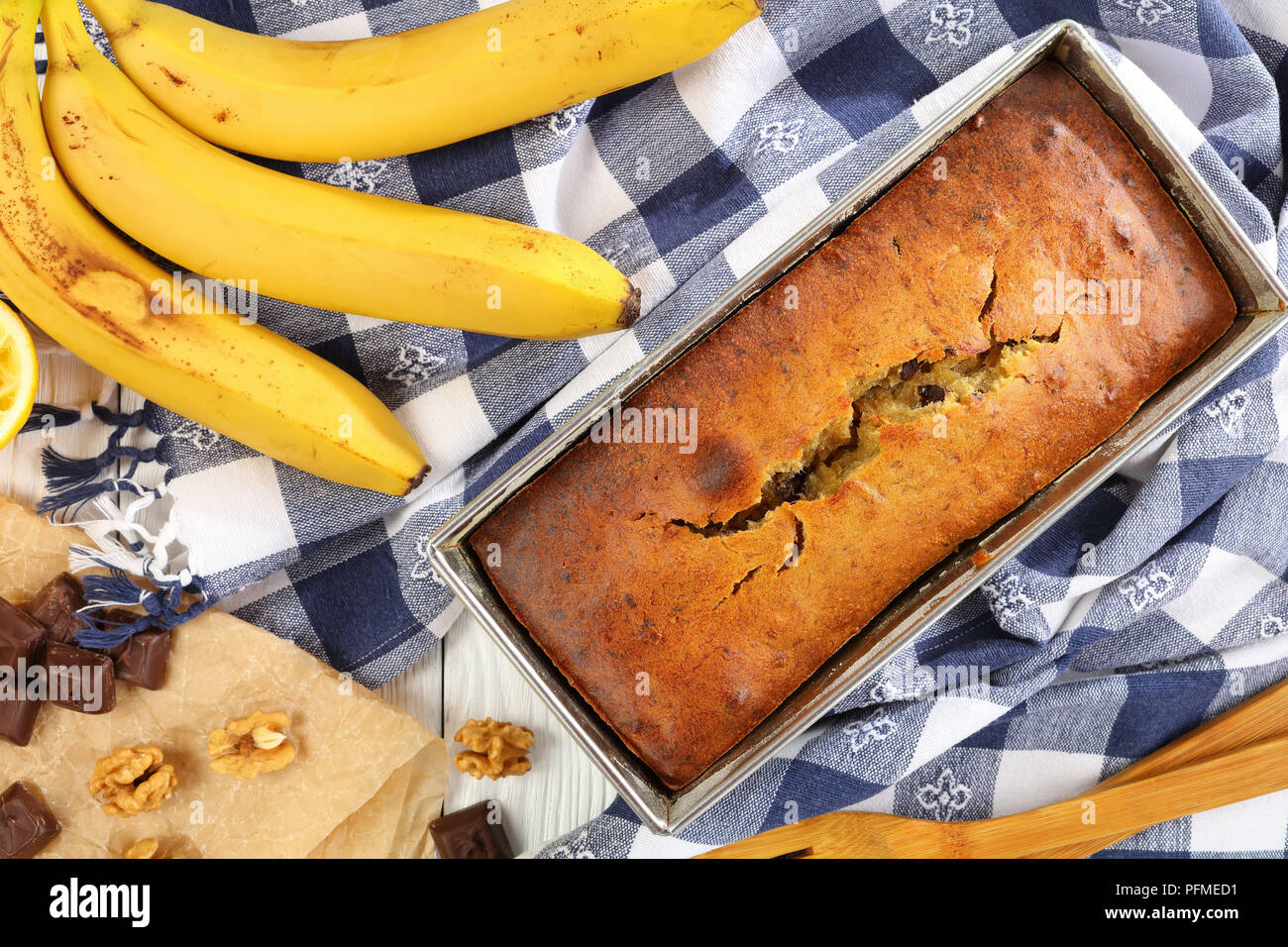freshly baked hot delicious banana bread with walnuts and chocolate pieces in a loaf pan with kitchen towel, ripe bananas and ingredients on wooden ta Stock Photo