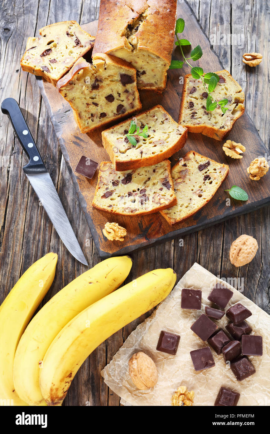 freshly baked delicious banana bread with walnuts and chocolate pieces cut in slices on cutting board. ingredients on rustic wooden table, vertical vi Stock Photo