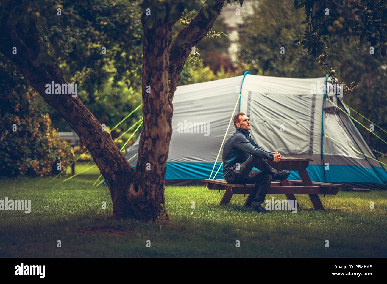 Summer Tent Camping Relax. Caucasian Men Relaxing on Wooden Bench in Front of His Tent. Campsite Theme. Stock Photo