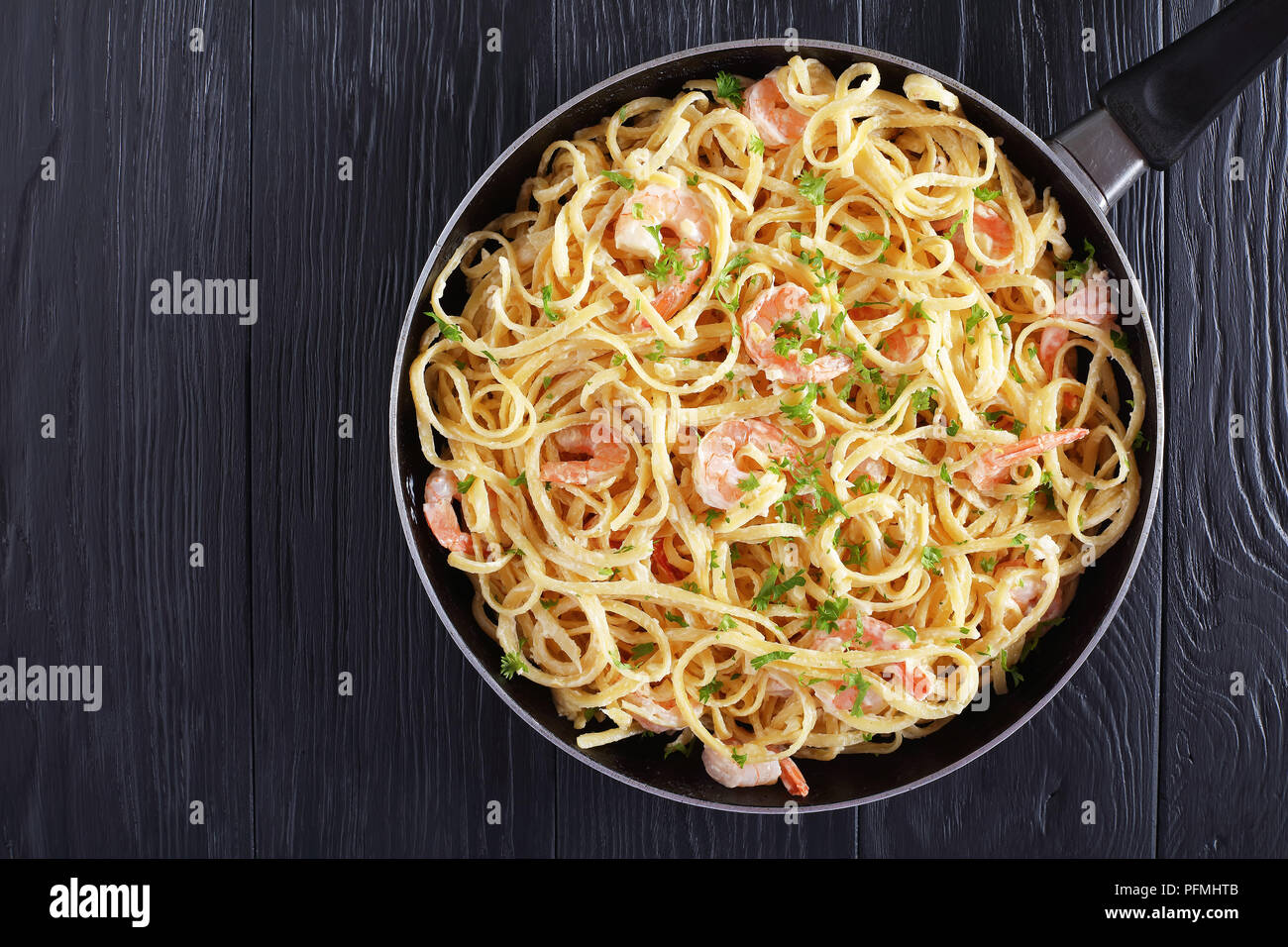Creamy Shrimp Alfredo linguine sprinkled with finely chopped parsley in skillet on black wooden table, easy recipe, view from above Stock Photo