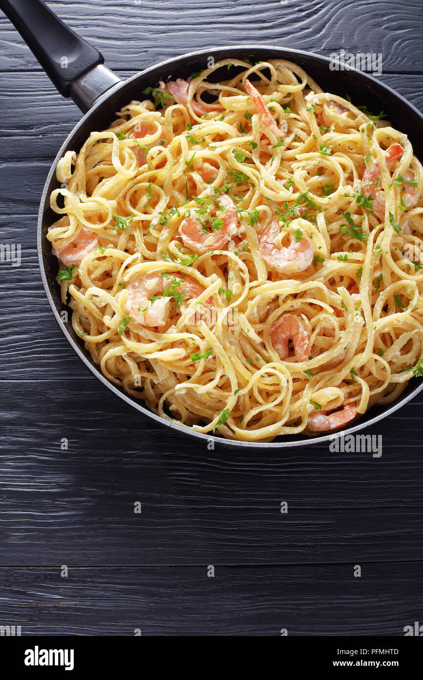 delicious Creamy Shrimp Alfredo linguine sprinkled with finely chopped parsley in skillet on black wooden table, vertical view from above, close-up Stock Photo