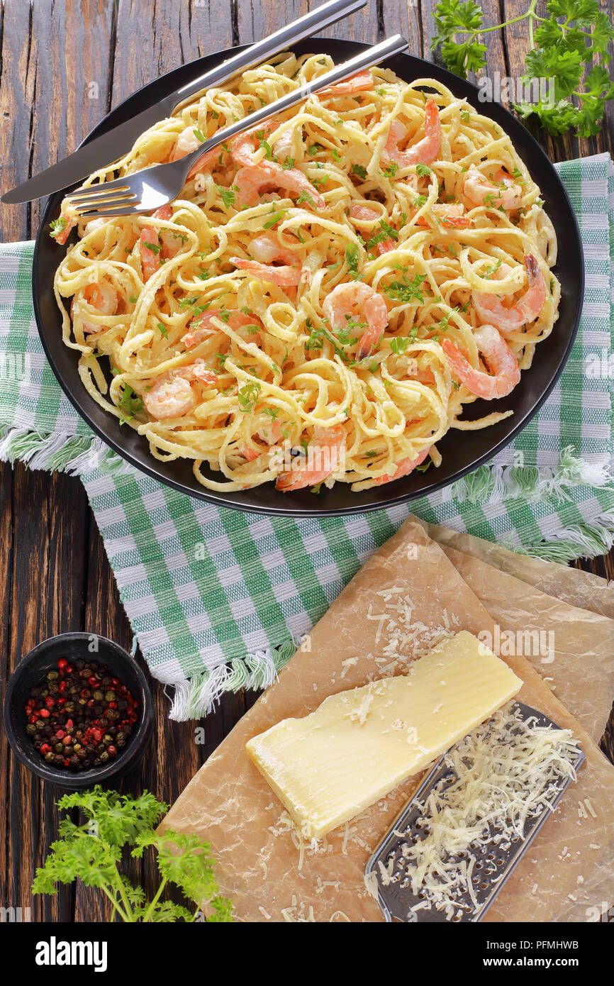 portion of Creamy Parmesan Shrimp pasta sprinkled with finely chopped parsley on black plate on wooden rustic table. piece of parmesan and grater at b Stock Photo