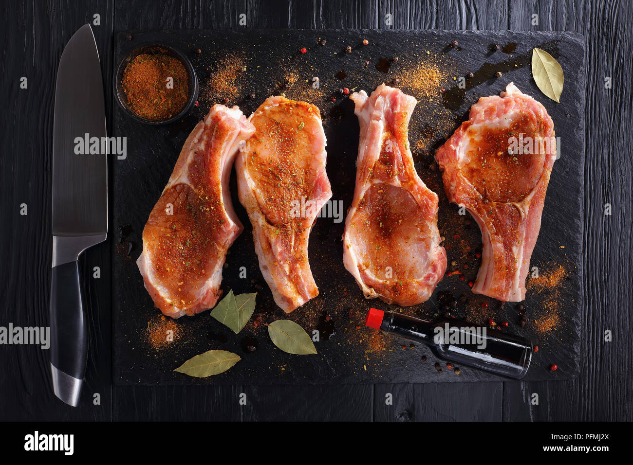 raw pork chops seasoned with spices, peppercorns and bay leaf on a black slate tray on wooden table with bottle of worcester sauce and knife, view fro Stock Photo