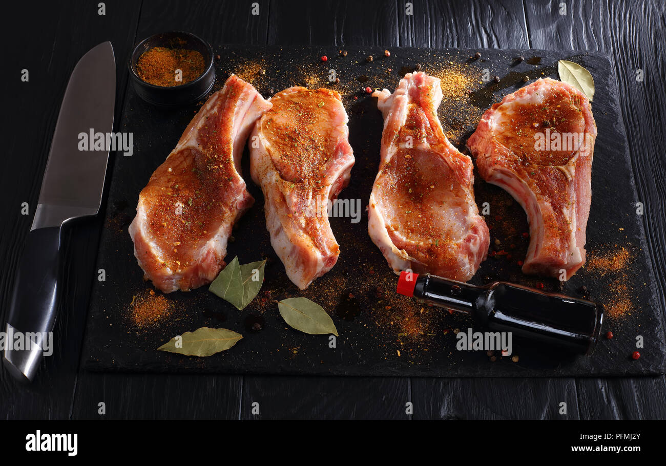 raw pork chops seasoned with spices, peppercorns and bay leaf on a black slate tray on wooden table with bottle of worcester sauce and knife, view fro Stock Photo