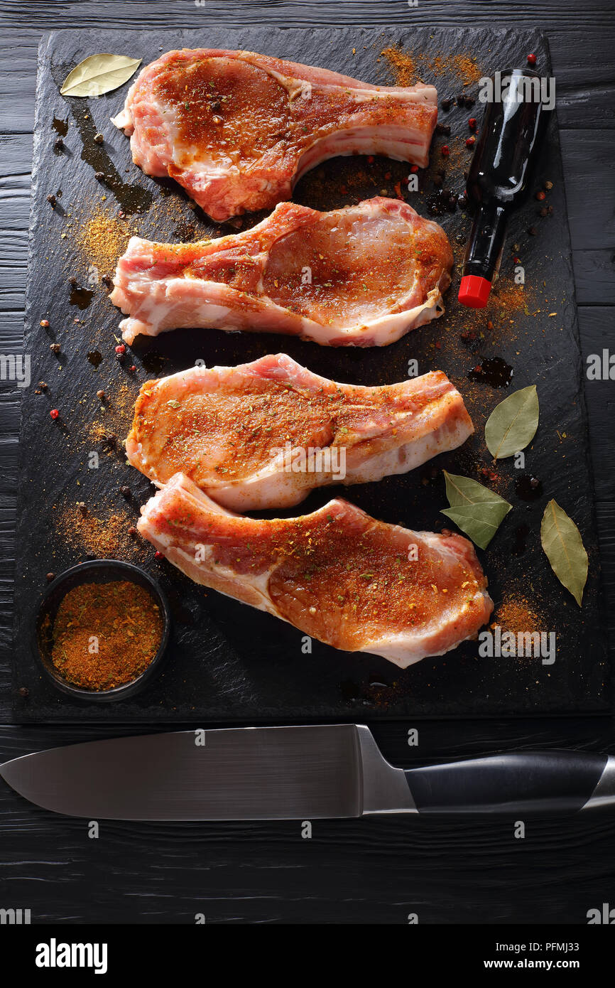 raw pork chops seasoned with spices, peppercorns and bay leaf on a black slate tray on wooden table with bottle of worcester sauce and knife, vertical Stock Photo