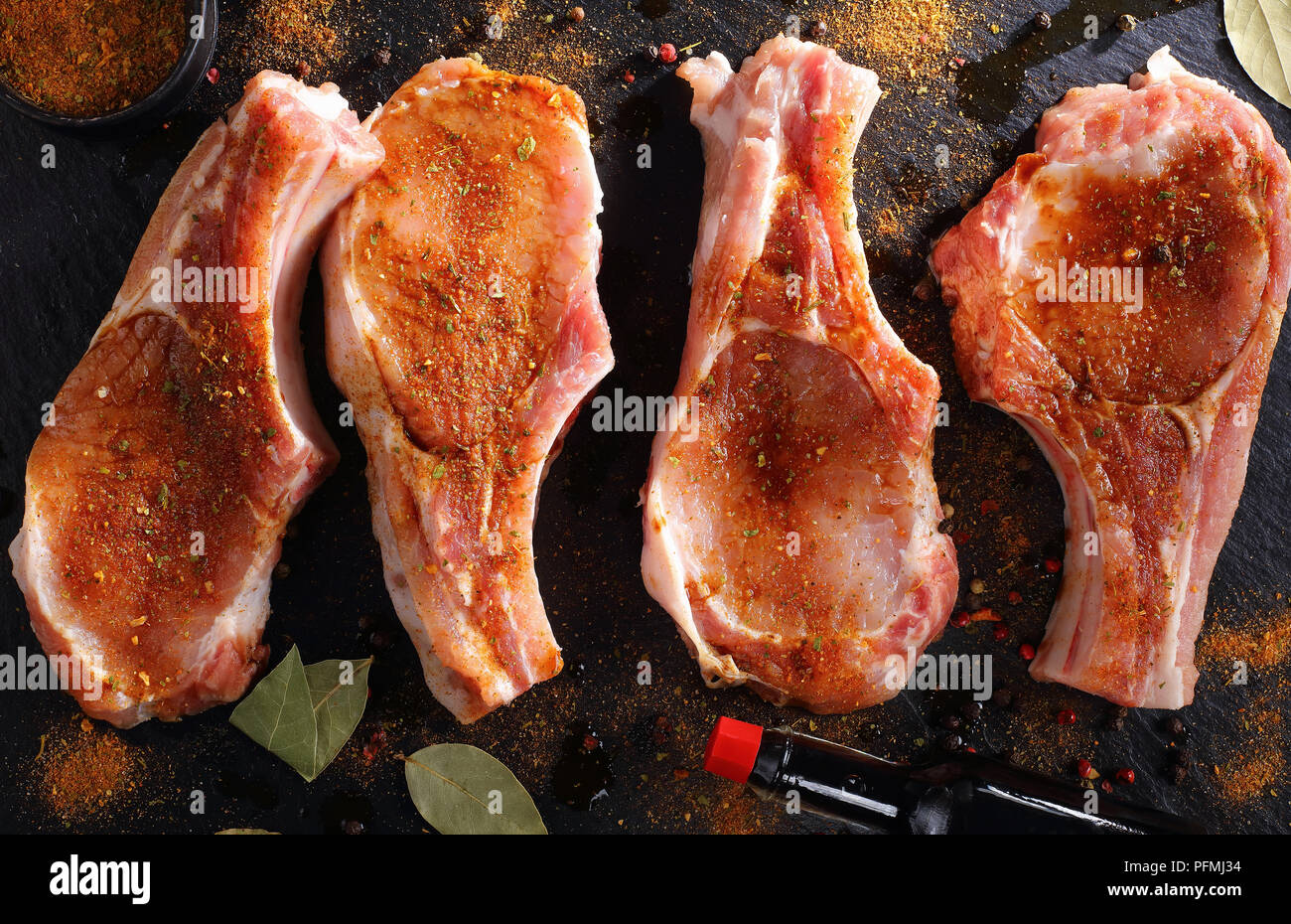 raw pork chops on bone seasoned with spices, peppercorns and bay leaf on a black slate tray on wooden table with bottle of worcester sauce and knife,  Stock Photo