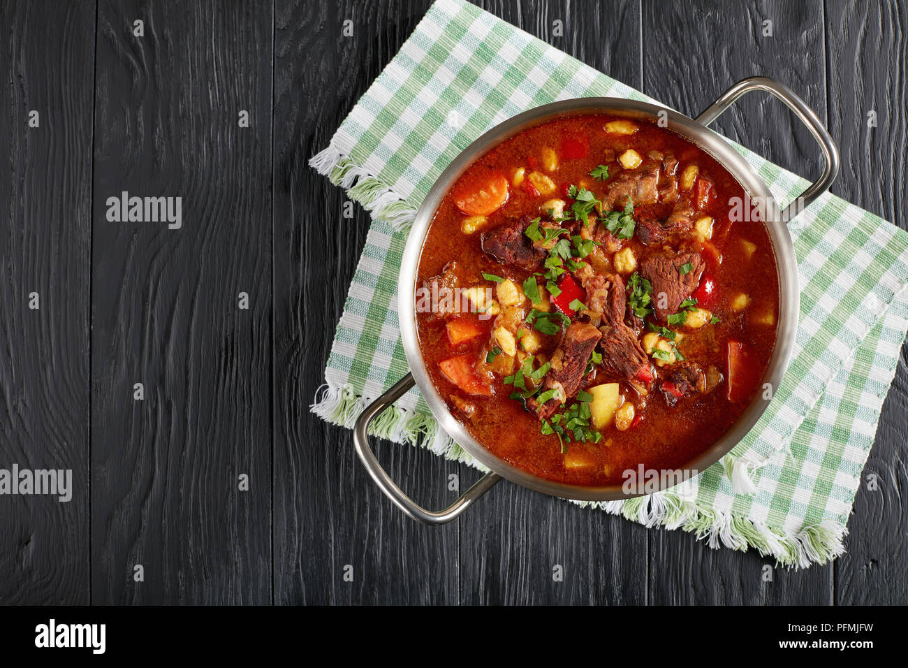delicious hot hungarian goulash with beef meat, paprika, vegetables and csipetke - small egg noodles in stainless steel pot on black wooden table, cla Stock Photo
